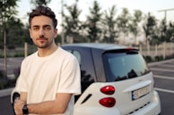A person with short hair and a beard wearing a white T-shirt and a smartwatch is standing in front of a small white car in a parking lot. Trees and parked cars are visible in the background.