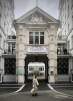 A person walks carrying several rolls of yellow material in front of a grand, ornate building with classical architectural details and a sign that reads 'Palmers Immobilien.' The structure has intricate molding and statues, with large windows and a passageway leading further inside.