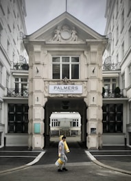 A person walks carrying several rolls of yellow material in front of a grand, ornate building with classical architectural details and a sign that reads 'Palmers Immobilien.' The structure has intricate molding and statues, with large windows and a passageway leading further inside.