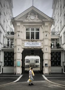 A person walks carrying several rolls of yellow material in front of a grand, ornate building with classical architectural details and a sign that reads 'Palmers Immobilien.' The structure has intricate molding and statues, with large windows and a passageway leading further inside.