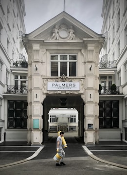 A person walks carrying several rolls of yellow material in front of a grand, ornate building with classical architectural details and a sign that reads 'Palmers Immobilien.' The structure has intricate molding and statues, with large windows and a passageway leading further inside.
