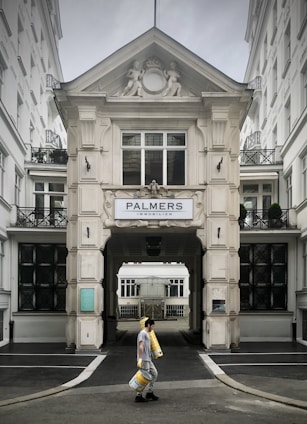 A person walks carrying several rolls of yellow material in front of a grand, ornate building with classical architectural details and a sign that reads 'Palmers Immobilien.' The structure has intricate molding and statues, with large windows and a passageway leading further inside.