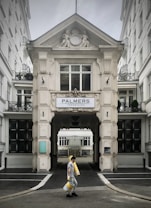 A person walks carrying several rolls of yellow material in front of a grand, ornate building with classical architectural details and a sign that reads 'Palmers Immobilien.' The structure has intricate molding and statues, with large windows and a passageway leading further inside.