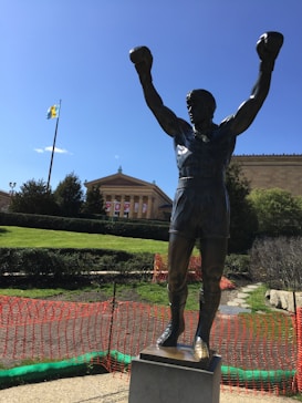 A bronze statue of a boxer with raised arms stands prominently in front of a classical-style building. The figure has its fists clenched and wears boxing shorts. In the background, a large flag flutters beside trees and a clear sky. The area around the statue is bordered with orange safety netting.