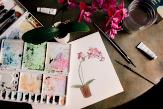 A bright studio table with watercolor paints, brushes, and a half-finished floral painting.