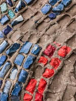 Colorful mosaic pattern featuring irregularly shaped chunks of vibrant blue and red stones embedded in a rough, gray-brown cement-like material, creating a textural contrast.