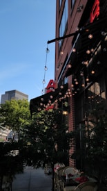 A cozy outdoor cafe setting with string lights hanging from a brick building, creating a warm and inviting atmosphere. The cafe has a small patio with tables and chairs partially obscured by green foliage. The sky is clear and blue, and a tall modern building is visible in the background.