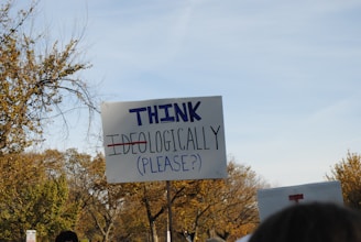 A protest sign reads 'THINK IDEOLOGICALLY (PLEASE?)' with the word 'IDEOLOGICALLY' crossed out. The scene includes trees with autumn foliage in the background, and there is another partially visible sign nearby. The sky is clear.