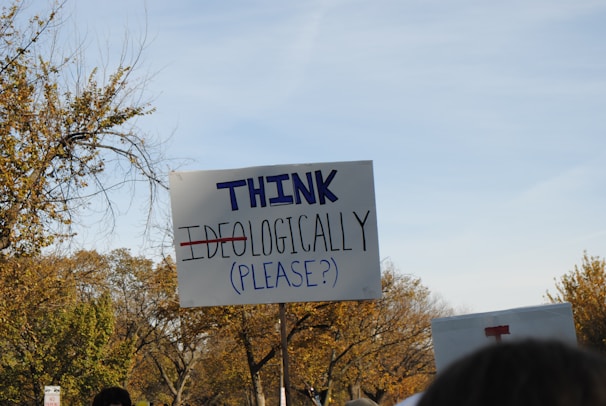 A protest sign reads 'THINK IDEOLOGICALLY (PLEASE?)' with the word 'IDEOLOGICALLY' crossed out. The scene includes trees with autumn foliage in the background, and there is another partially visible sign nearby. The sky is clear.
