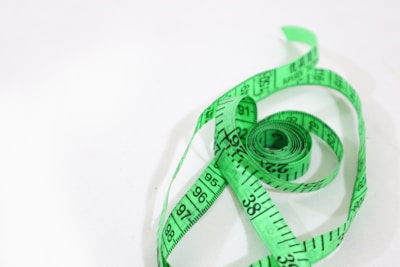 A coiled green measuring tape is placed on a white surface. The tape is partially unrolled, displaying numerical measurements in black print.
