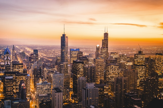 A bustling cityscape featuring a skyline of towering skyscrapers under a vibrant sunset. The buildings are illuminated with lights, creating a lively and energetic atmosphere as the city transitions from day to night.