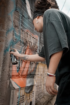 A person with dreadlocks, wearing a black headband, large earring, and a dark t-shirt, is spray painting a mural on a rough, brick wall. The wall features vibrant, multicolored graffiti with shades of blue, orange, and white. The person is holding a black spray paint can, concentrating on their artistic work. They also have visible tattoos on their arm and a bracelet on their wrist.