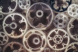 A series of overlapping metallic gears, intricately arranged, with varying sizes and shapes. The aged texture of the metal suggests a vintage or steampunk aesthetic.