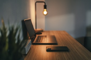 A wooden desk holds a closed laptop, a smartphone, and a small speaker. A modern desk lamp with an exposed bulb casts a warm glow, creating a cozy and focused workspace. The background appears softly blurred, with hints of greenery.