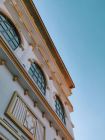 An upward angle view of an elegant building featuring arches and large windows, adorned with decorative architectural elements in a beige and white color palette against a clear blue sky.