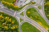 An aerial view of a large, complex road intersection featuring multiple lanes, a roundabout, and adjacent parking lot. Surrounding the roads are dense green areas with trees and grass, as well as open space with sparse vegetation. Vehicles are visible on the roads and parked in the lot.