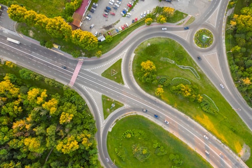 An aerial view of a large, complex road intersection featuring multiple lanes, a roundabout, and adjacent parking lot. Surrounding the roads are dense green areas with trees and grass, as well as open space with sparse vegetation. Vehicles are visible on the roads and parked in the lot.