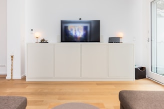 A modern living room with a sleek, white cabinet set against a white wall. A flat-screen TV is mounted on top of the cabinet, flanked by two small table lamps with round white shades. The room has wooden flooring and there's a large window or glass door on the right side, allowing natural light to enter. A decorative piece resembling a twisted candle holder is on the floor to the left of the cabinet.