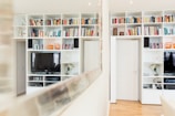 A modern living room featuring white built-in shelving units filled with colorful books, decorative items, and two televisions. The room has polished wooden flooring and is well-lit with recessed ceiling lights. A large mirror reflects part of the bookcase and room.