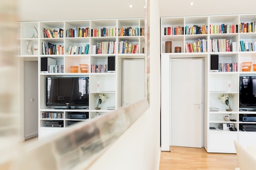 A modern living room featuring white built-in shelving units filled with colorful books, decorative items, and two televisions. The room has polished wooden flooring and is well-lit with recessed ceiling lights. A large mirror reflects part of the bookcase and room.