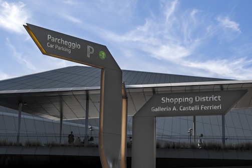 A modern architectural structure with a detailed roof design stands in the background. In the foreground, two directional signs point to different locations: one leading to a parking area labeled 'Parcheggio Car Parking' with a parking symbol and another directing to a shopping district named 'Galleria A. Castelli Ferrieri.' The sky above is clear with a few wispy clouds scattered across.