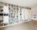 A spacious room with a large built-in bookshelf covering the entire wall, filled with various books, records, and decorative items. The shelves are predominantly white, creating a clean and modern look. The floor is wooden, and a couple of framed pictures are hung on the adjacent wall. Some shelves hold electronic devices and some have closed doors.