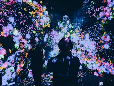A vibrant display of colorful floral projections illuminates a dark room, creating an immersive art experience. Several individuals are present, interacting and observing the captivating digital blossoms scattered across the walls and floor.