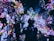 A vibrant display of colorful floral projections illuminates a dark room, creating an immersive art experience. Several individuals are present, interacting and observing the captivating digital blossoms scattered across the walls and floor.