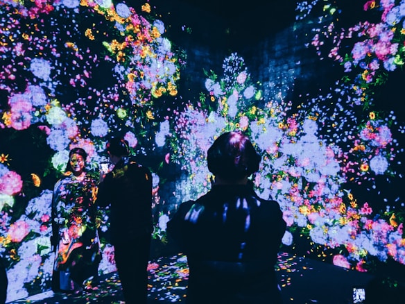 A vibrant display of colorful floral projections illuminates a dark room, creating an immersive art experience. Several individuals are present, interacting and observing the captivating digital blossoms scattered across the walls and floor.