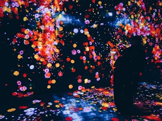 A vibrant, immersive digital art installation featuring a multitude of colorful flowers projected onto walls and the floor. The scene is dark, allowing the bright colors of the flowers, including reds, yellows, and pinks, to stand out. Two individuals are present, silhouetted against the colorful backdrop, seemingly engaged with their surroundings.