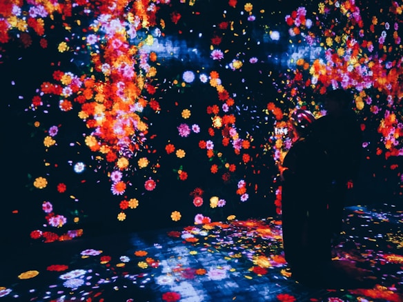A vibrant, immersive digital art installation featuring a multitude of colorful flowers projected onto walls and the floor. The scene is dark, allowing the bright colors of the flowers, including reds, yellows, and pinks, to stand out. Two individuals are present, silhouetted against the colorful backdrop, seemingly engaged with their surroundings.