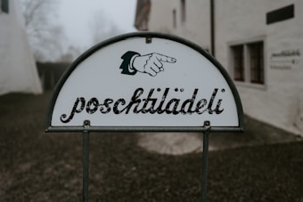 A sign with the word 'poschtilädeli' displayed in stylized script, accompanied by an illustration of a pointing hand. The background features blurred buildings and trees, creating a subdued and misty atmosphere.
