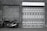 A black motorcycle is parked next to a wall with a brick texture and a large garage door. The garage door is decorated with various geometric patterns and images of rabbits. The scene is captured in black and white, giving it a vintage feel.