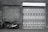 A black motorcycle is parked next to a wall with a brick texture and a large garage door. The garage door is decorated with various geometric patterns and images of rabbits. The scene is captured in black and white, giving it a vintage feel.