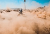 A person is riding a quad bike on a dirt path, creating a large cloud of dust in the air. The scene is set in an open, rugged landscape with dry grass and distant hills visible under a clear blue sky. The atmosphere conveys a sense of action and adventure.