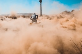 A person is riding a quad bike on a dirt path, creating a large cloud of dust in the air. The scene is set in an open, rugged landscape with dry grass and distant hills visible under a clear blue sky. The atmosphere conveys a sense of action and adventure.