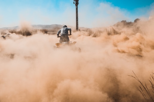 A person is riding a quad bike on a dirt path, creating a large cloud of dust in the air. The scene is set in an open, rugged landscape with dry grass and distant hills visible under a clear blue sky. The atmosphere conveys a sense of action and adventure.