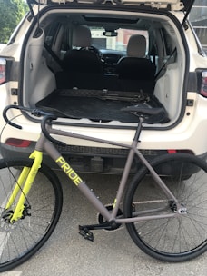 A bicycle is positioned behind a parked white SUV with its rear hatch open. The bike is branded 'PRIDE' and is primarily gray with bright yellow accents on the fork. The SUV's interior is visible, showing folded seats and a spacious cargo area.