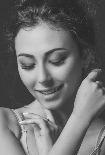A close-up portrait of a woman with eyes closed, smiling gently. There is a sense of serenity and contentment in her expression. Her hands are gracefully positioned near her face, showcasing delicate rings on her fingers.