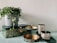 A collection of decorative items is arranged on a surface. A grey pot with trailing green foliage sits atop a rectangular box with intricate green and gold patterns. A two-tiered tray with gold interiors and green accents adds to the arrangement. Nearby, two cylindrical containers, decorated with black leaf designs, are placed on a matching green tray.