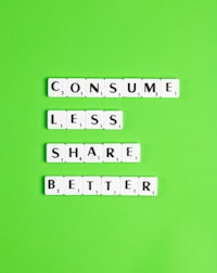 Scrabble tiles arranged on a bright green background spell out the words 'CONSUME LESS SHARE BETTER.' The letters are neatly aligned in three horizontal lines.