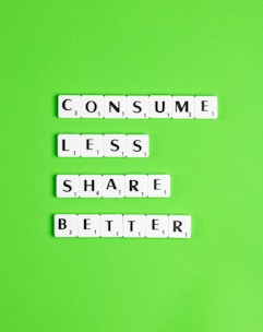 Scrabble tiles arranged on a bright green background spell out the words 'CONSUME LESS SHARE BETTER.' The letters are neatly aligned in three horizontal lines.