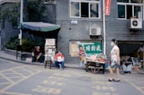 A street scene with a small makeshift market setup outside a tiled building. Two people are seated near tables displaying various fruits such as watermelons and grapes. A woman in a floral top and shorts walks by, wearing a face mask. The street is marked with yellow painted lines, and signs are visible on the building's exterior, including one with Chinese characters.