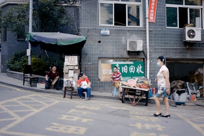 A street scene with a small makeshift market setup outside a tiled building. Two people are seated near tables displaying various fruits such as watermelons and grapes. A woman in a floral top and shorts walks by, wearing a face mask. The street is marked with yellow painted lines, and signs are visible on the building's exterior, including one with Chinese characters.