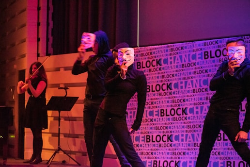 Several individuals dressed in black attire are performing on a stage. They are holding white masks in front of their faces, adding an element of mystery. The backdrop features the word 'BLOCKCHANCE' repeatedly. One person is playing a violin.