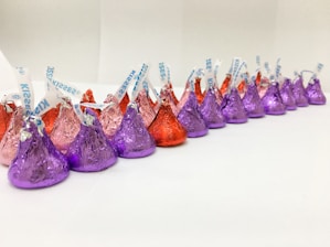 A neatly arranged line of colorful, foil-wrapped, teardrop-shaped chocolates. The chocolates are wrapped in foil of purple, red, and pink hues, each with a small paper strip poking out from the top.
