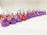 A neatly arranged line of colorful, foil-wrapped, teardrop-shaped chocolates. The chocolates are wrapped in foil of purple, red, and pink hues, each with a small paper strip poking out from the top.