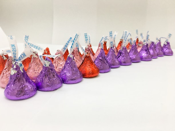 A neatly arranged line of colorful, foil-wrapped, teardrop-shaped chocolates. The chocolates are wrapped in foil of purple, red, and pink hues, each with a small paper strip poking out from the top.