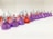 A neatly arranged line of colorful, foil-wrapped, teardrop-shaped chocolates. The chocolates are wrapped in foil of purple, red, and pink hues, each with a small paper strip poking out from the top.