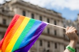 A hand holding a rainbow-colored flag, indicative of LGBTQ+ pride, in front of a blurred building. The flag features horizontal stripes in red, orange, yellow, green, blue, and purple.